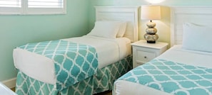 A room with two beds, each with a white headboard and a teal patterned bedspread. There is a nightstand with a lamp between the beds.