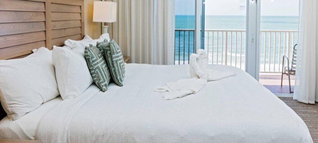 Habitación de hotel con una cama grande, ropa de cama blanca, cabecera de madera, mesita de noche con lámpara y vista al océano desde un balcón.