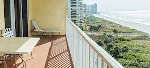 A balcony with a table and two chairs, overlooking a beach and a coastal road.