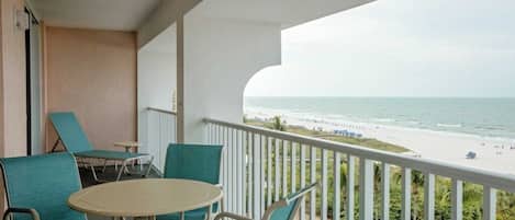 A balcony with a view of the beach, a table, and two chairs.