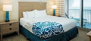 A hotel room with a bed, two bedside tables, a nightstand, a window with a view of the ocean, and a balcony with a chair.