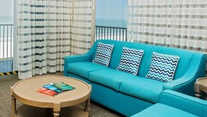 Een kamer met een blauwe bank, gestreepte gordijnen, een ronde houten salontafel en uitzicht op het strand.