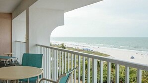 A balcony with a view of the beach, a table, and two chairs.