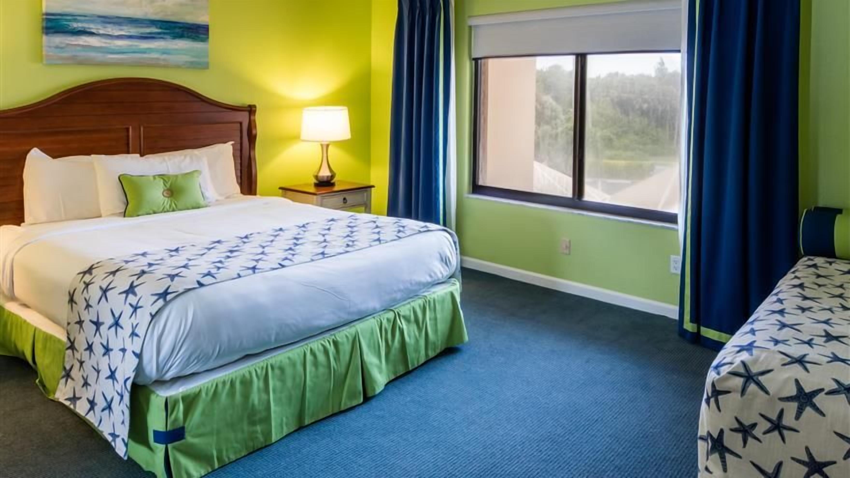 A hotel room with a large bed, a nightstand, a window with curtains, and a view of the outdoors.