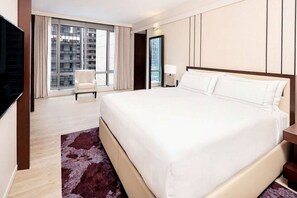 Habitación de hotel con una cama grande, televisión, silla, escritorio y vista a un edificio por la ventana.