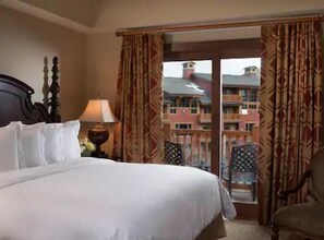 A hotel room with a bed, a chair, a nightstand, and a view of a building through the window.