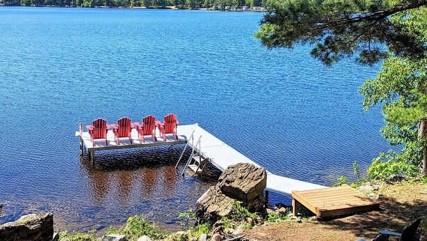 A lakeside dock with red chairs and a wooden table, surrounded by natural scenery and trees.