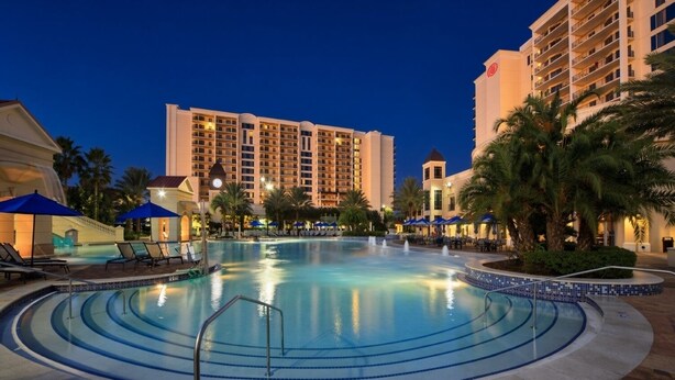 A hotel pool area with multiple lounge chairs and umbrellas, surrounded by palm trees and a large hotel building in the background. - Orlando | Romantic Park Retreat | Pool + Parks (Orlando)