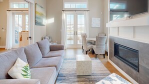 A living room with a grey sofa, a coffee table, and a fireplace. There are two windows with curtains, a dining area with chairs, and a view of the outside. - Resort Pools, Golf Cart Boardwalk to Beach (Port Aransas)