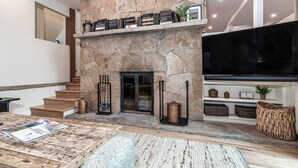 A modern living room with a stone fireplace, a staircase, a flat-screen TV, and a coffee table with magazines.