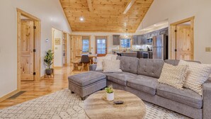 A spacious living room with a large gray sectional sofa, wooden ceiling beams, and a kitchen area in the background. - Ellijay Escape: Hot Tub, Fireplace, Peaceful Views (Ellijay)