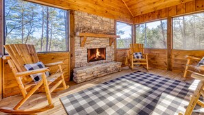A log cabin interior with a stone fireplace, wooden rocking chairs, and large windows offering a forest view. - Ellijay Escape: Hot Tub, Fireplace, Peaceful Views (Ellijay)
