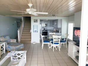 Un salon avec un canapé, une table à manger avec des chaises, un téléviseur, un coin cuisine avec des placards, un ventilateur de plafond et un escalier.