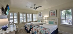 A bedroom with two beds, a desk with a computer, a ceiling fan, and large windows with blinds.