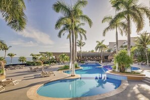 Ein Poolbereich eines Resorts mit Palmen, Liegestühlen und Blick aufs Meer.