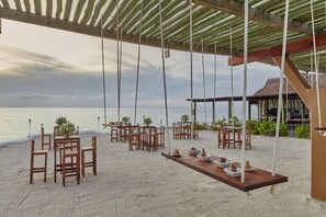 A beachside dining area with wooden tables and chairs, a swing, and a view of the ocean.