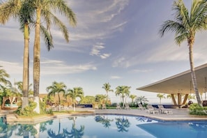 Zona de piscina con palmeras, sillones de descanso y un área de estar techada.
