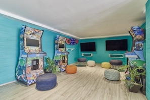 Ein Zimmer mit türkisfarbenen Wänden, zwei Spielautomaten, einem Flachbildfernseher und mehreren Hocker.