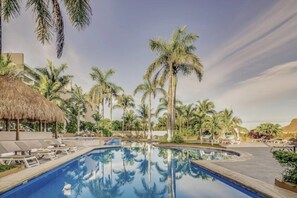 Ein Swimmingpool, umgeben von Palmen und Liegestühlen, mit einer strohgedeckten Struktur und blauem Himmel.