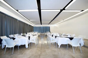 Un salón de conferencias con mesas y sillas blancas, una mesa larga en el centro, y un telón de fondo azul.