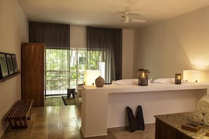 A modern hotel room with a large window, a ceiling fan, a wooden bench, a small table with towels, and a white reception desk.