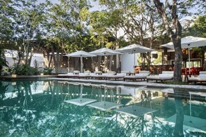 Área da piscina com espreguiçadeiras e guarda-sóis, cercada por árvores e um edifício com grandes janelas.