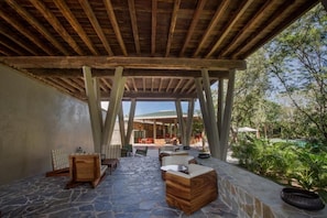 An outdoor patio with stone flooring, wooden furniture, and a view of a pool and greenery.