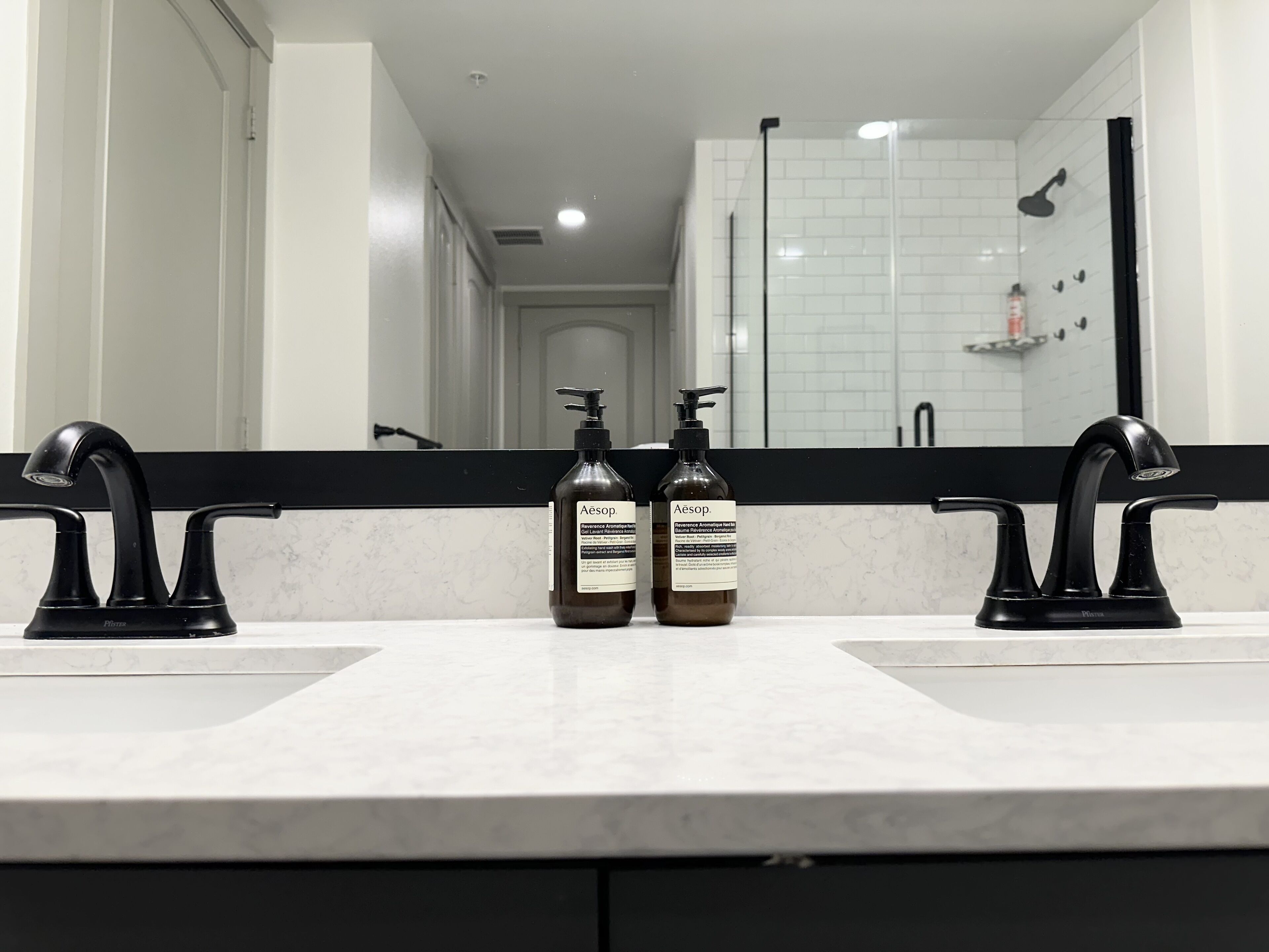 Casa de banho com duas torneiras pretas, bancada de mármore branco e dois frascos de shampoo Aesop.
