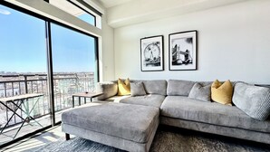 A modern living room with a large sectional sofa, a small side table, and two framed pictures on the wall. - The Heights Cozy Stay - Gym&pool 12mins to Midtown and Downtown (Houston)
