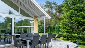 A modern outdoor dining area with a glass enclosure, a wooden deck, and a view of the surrounding greenery.