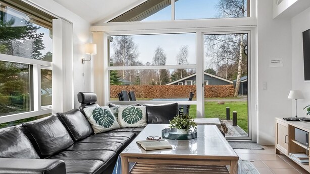 A modern living room with a leather sofa, a glass coffee table, and a view of a backyard through large windows.