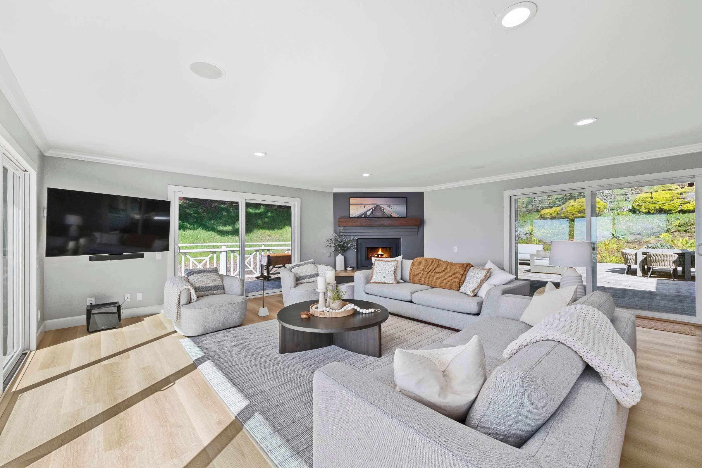 A modern living room with a grey sofa set, a flat-screen TV, a fireplace, and a view of a garden through large windows.