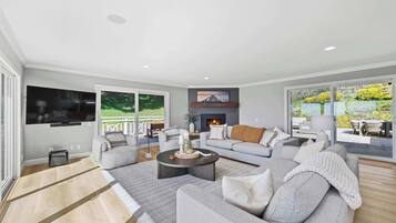 A modern living room with a grey sofa set, a flat-screen TV, a fireplace, and a view of a garden through large windows.