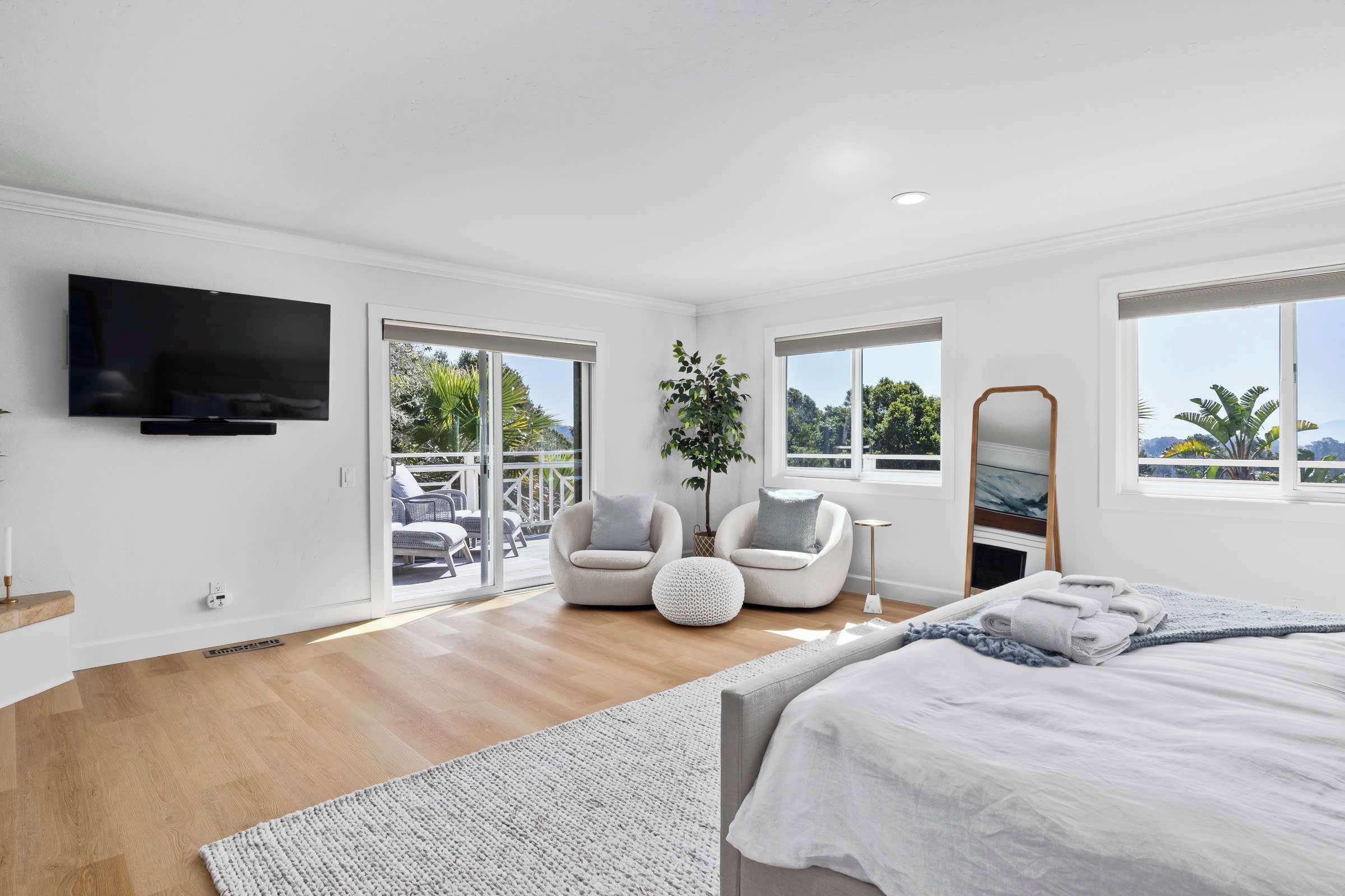 A modern bedroom with a flat-screen TV, a bed, a seating area with chairs, and large windows offering a view of greenery.