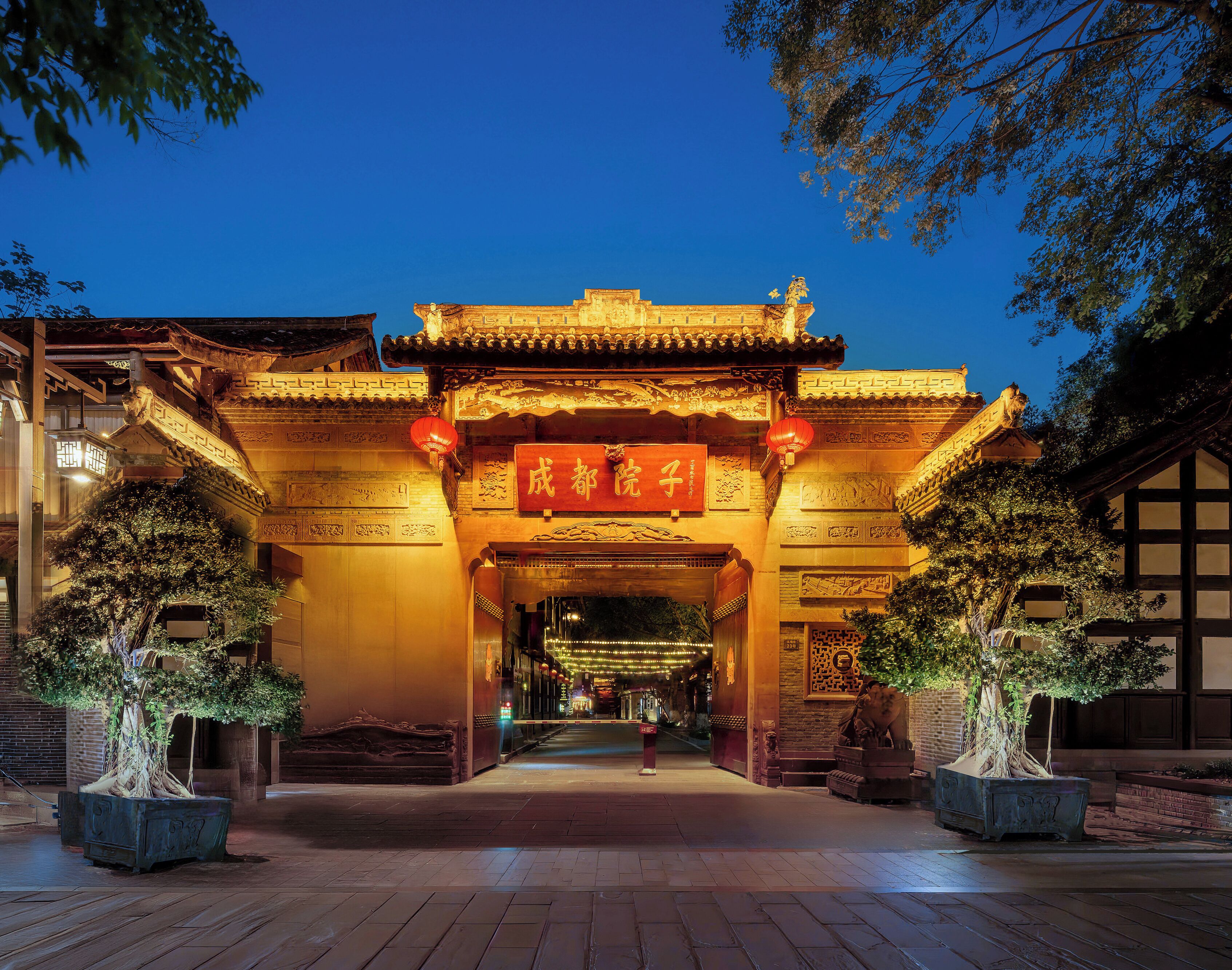 A traditional Chinese gate with intricate carvings and a red plaque, flanked by potted trees and illuminated by warm lighting.