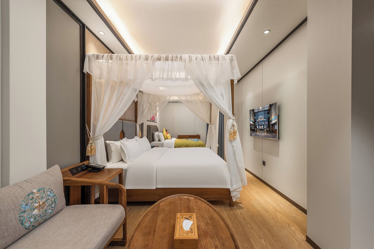 A modern hotel room with a bed, a sofa, a wooden side table, and a TV on the wall.