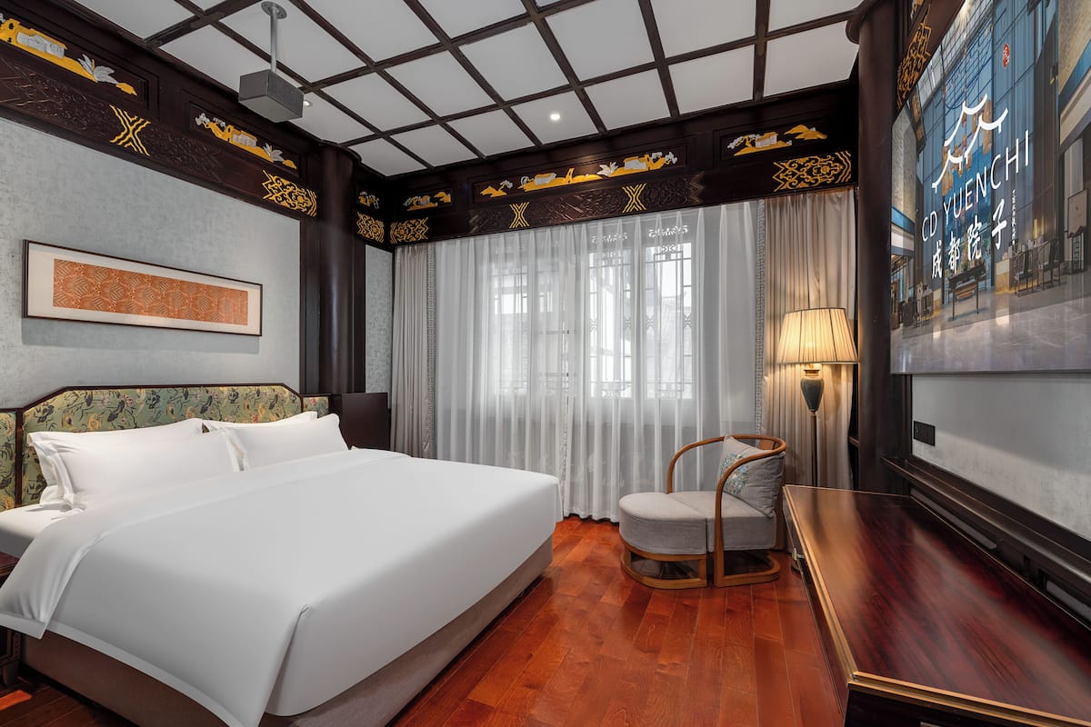 A hotel room with a large bed, a chair, a desk, and a large window with curtains.
