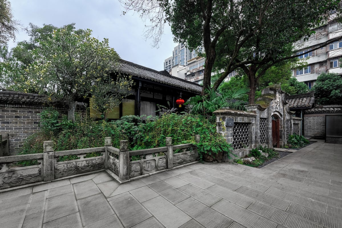 A traditional courtyard with a thatched-roof building, a stone fence with carvings, and lush greenery.