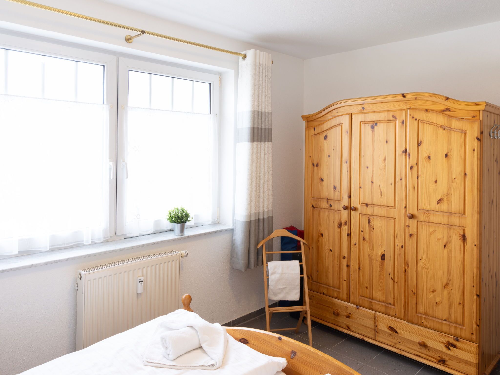 A bedroom with a bed, a wooden wardrobe, a radiator, a window with curtains, and a small potted plant on the windowsill.