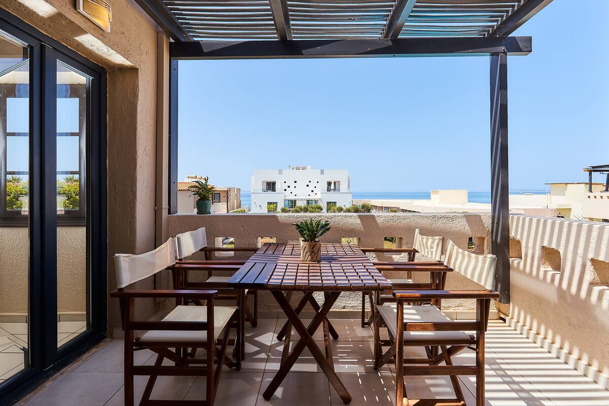 A terrace with a wooden table and chairs, a potted plant, and a view of the sea and buildings.
