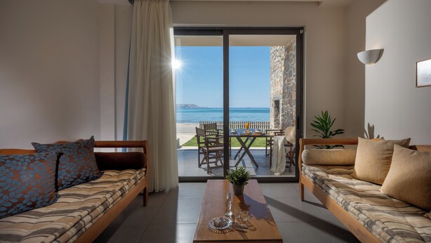A living room with a sofa, a coffee table, and a view of the sea through a glass door. - Creteco Hotel & Suites (Hersonissos)