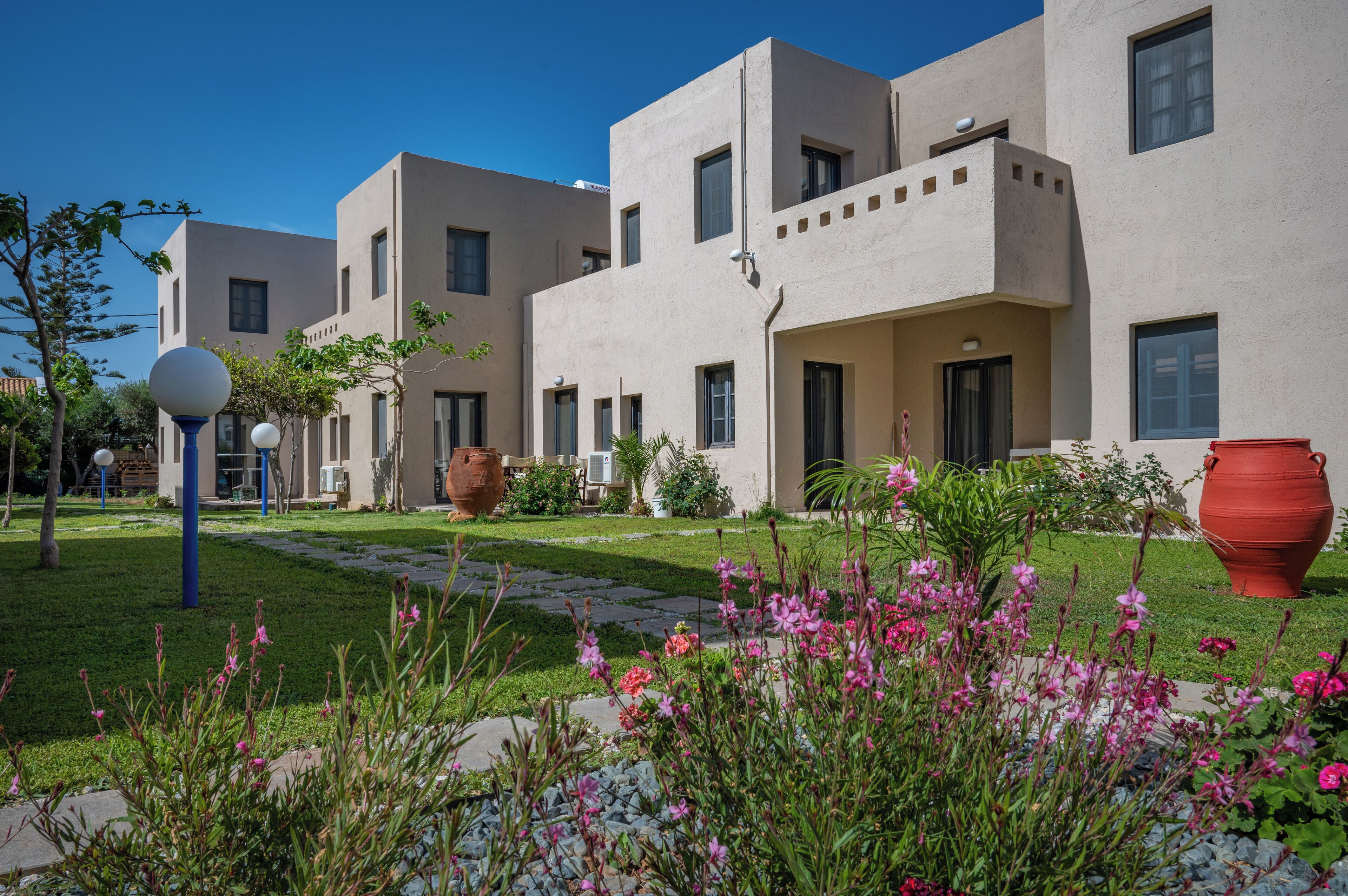 A modern building with a flat facade, multiple windows, and a small garden area with pink flowers and a red pot.