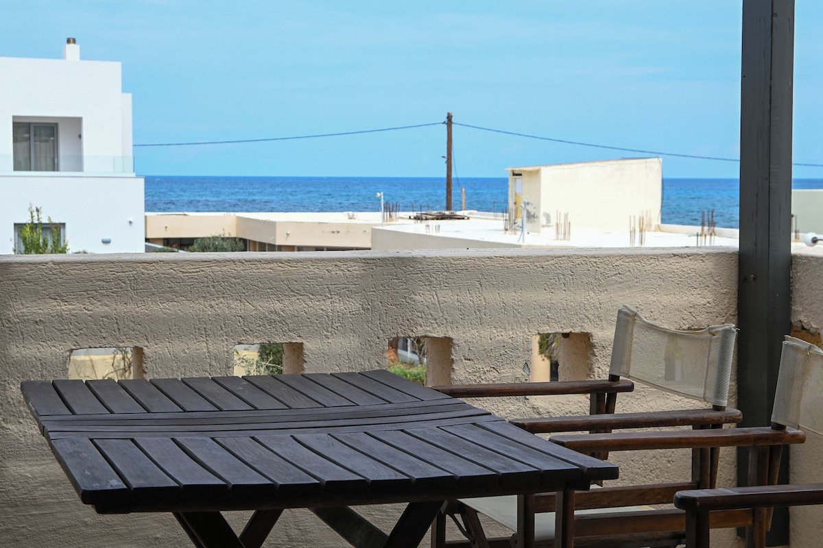 A terrace with a wooden table and benches, overlooking a coastal area with buildings and the sea.