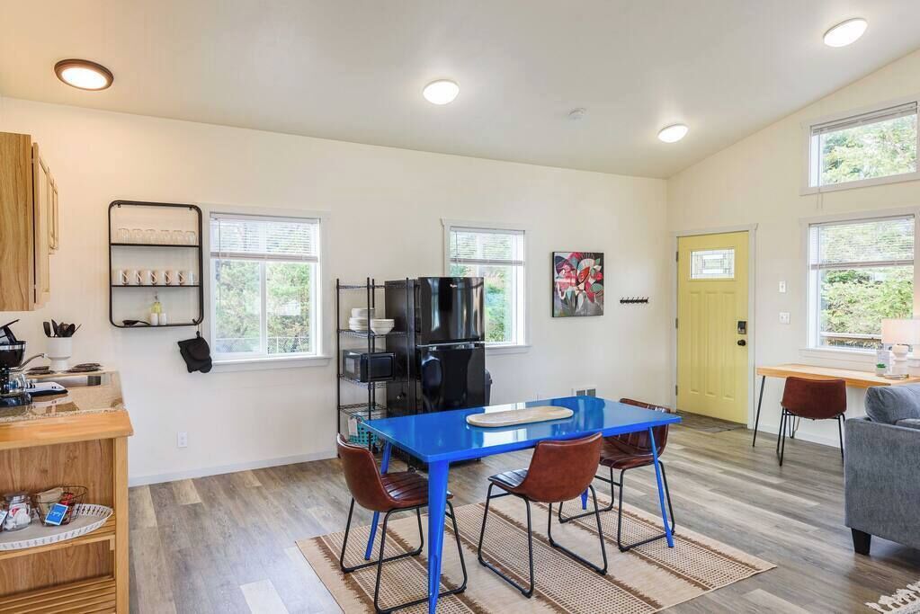 A modern kitchen with a blue table, two chairs, and a refrigerator. There are windows and a yellow door.