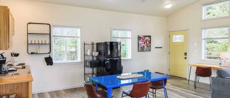 A modern kitchen with a blue table, two chairs, and a refrigerator. There are windows and a yellow door.