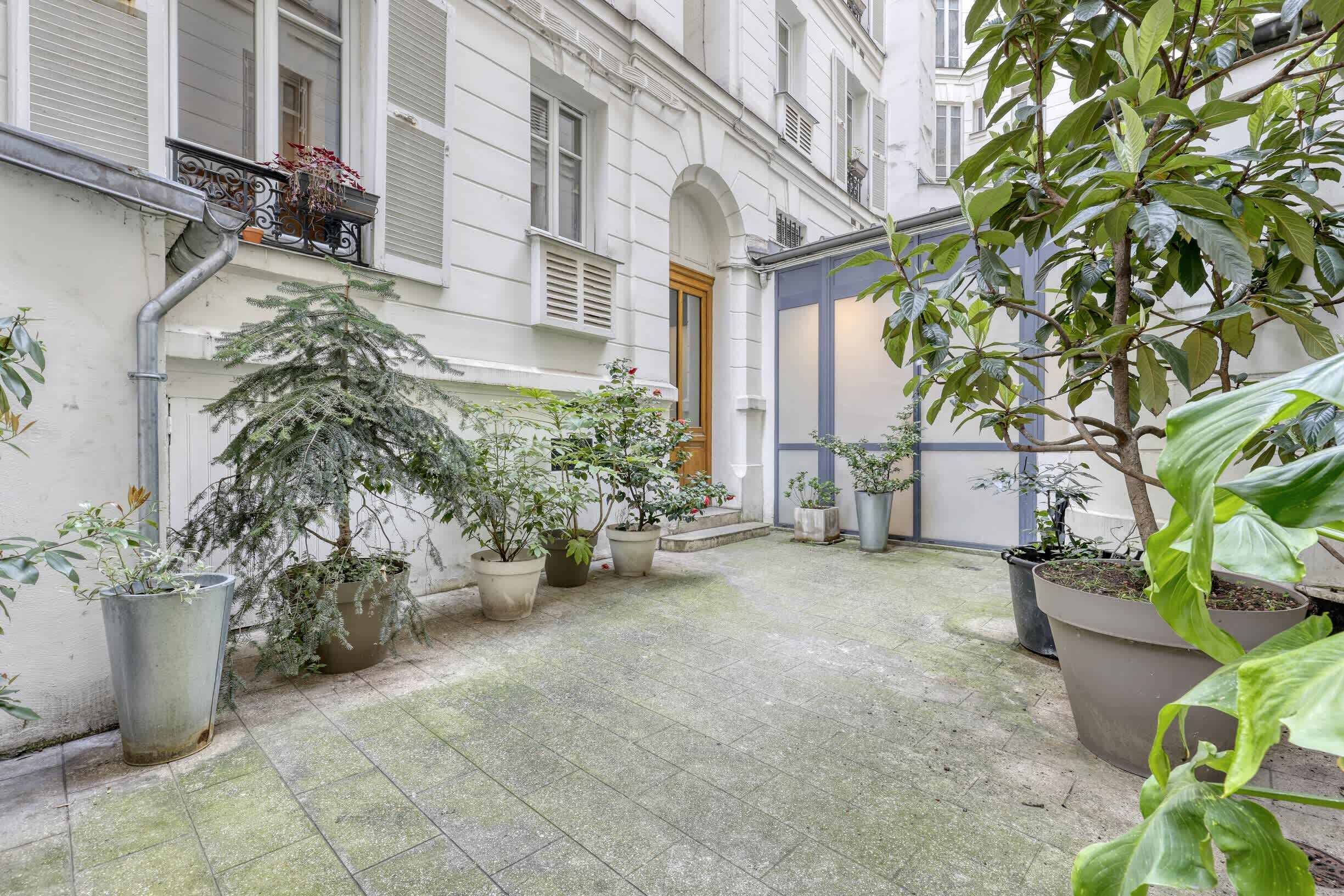 Un patio con plantas en macetas, piso de baldosas y un edificio de paredes blancas y puerta de madera.