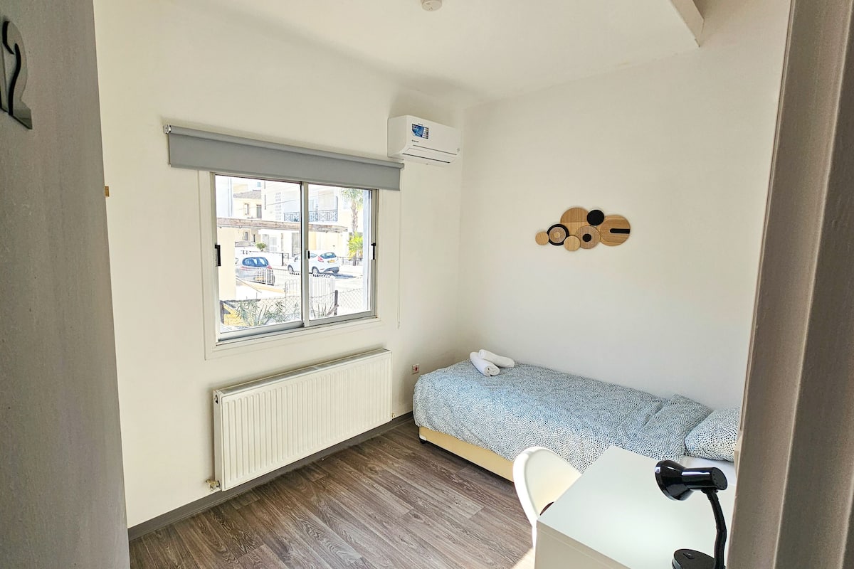 A small room with a bed, a radiator, a window with a view of the street, and a wall decoration.