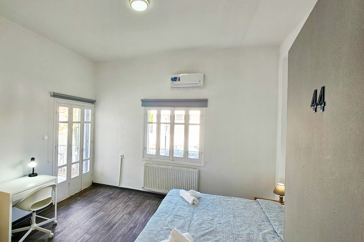 A small, well-lit room with a desk, a bed, and two windows.
