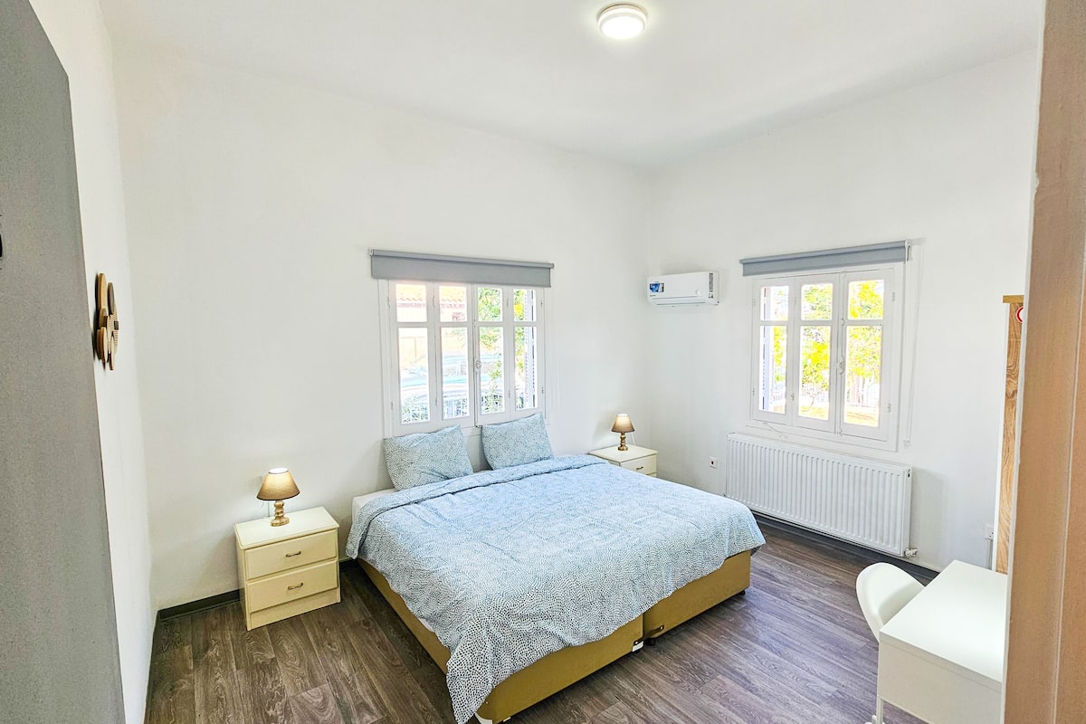 A bedroom with a bed, two bedside tables, a nightstand, a radiator, and two windows.