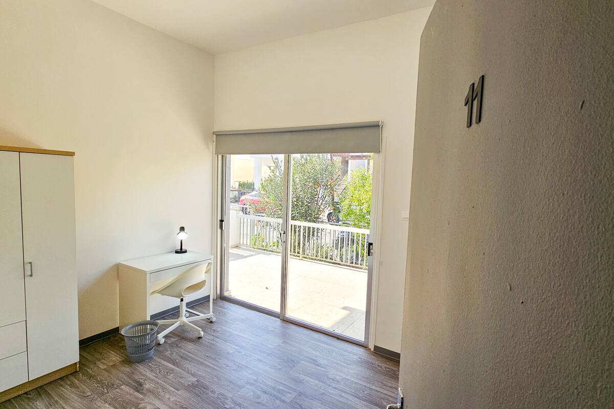 A room with a sliding glass door leading to a balcony, a desk with a lamp, and a small basket on the floor.